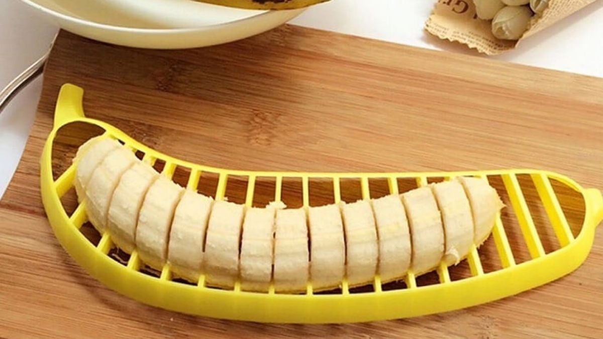 A peeled banana sits on a wooden cutting board, sliced evenly using a yellow banana-shaped slicer. The slices remain aligned within the slicer, and a plate with fruit is visible in the background.