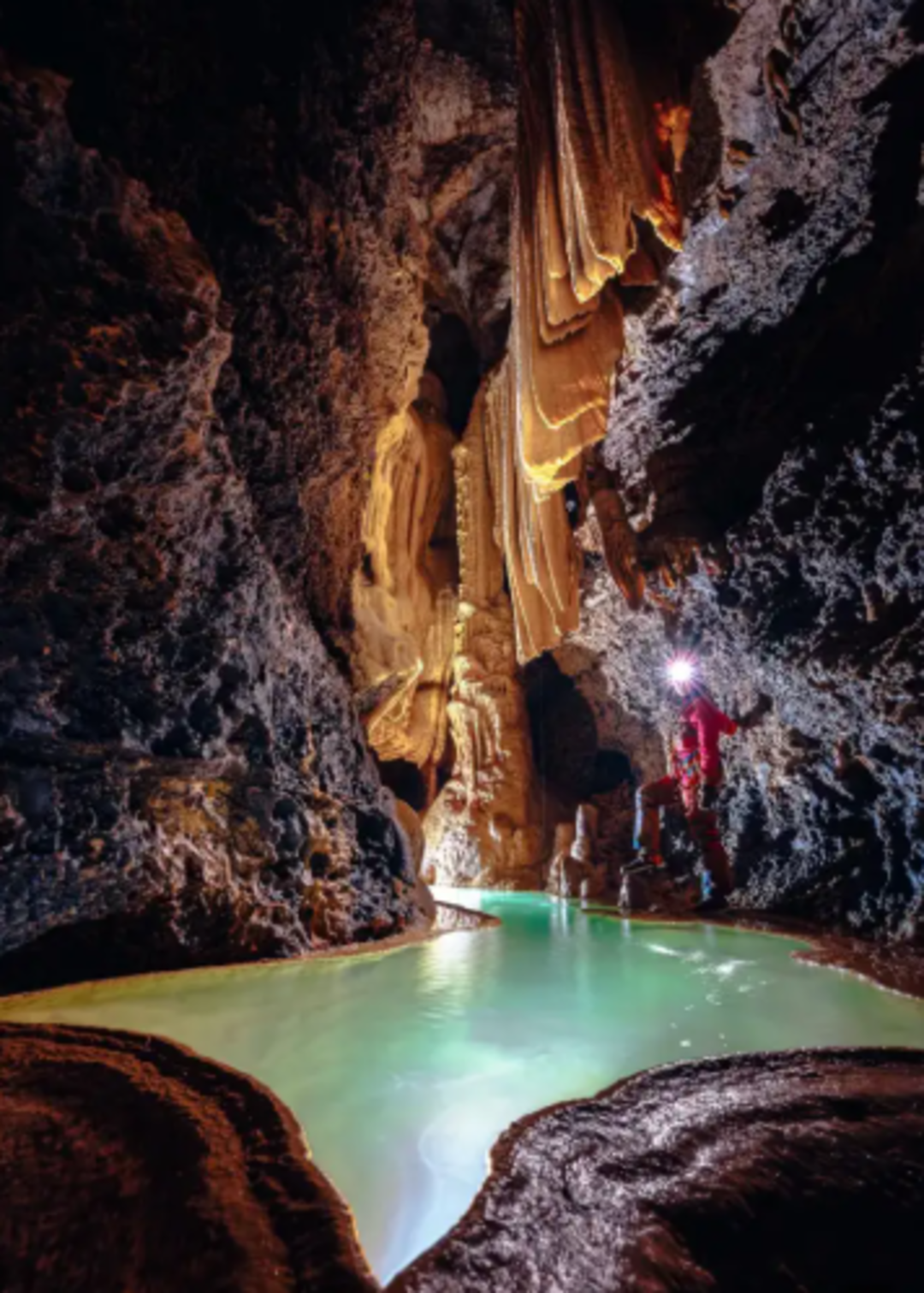 A person in red gear explores a striking cave with dramatic rock formations and a glowing turquoise pool of water, illuminated by a flashlight. Stalactites hang from the ceiling above the winding waterway.