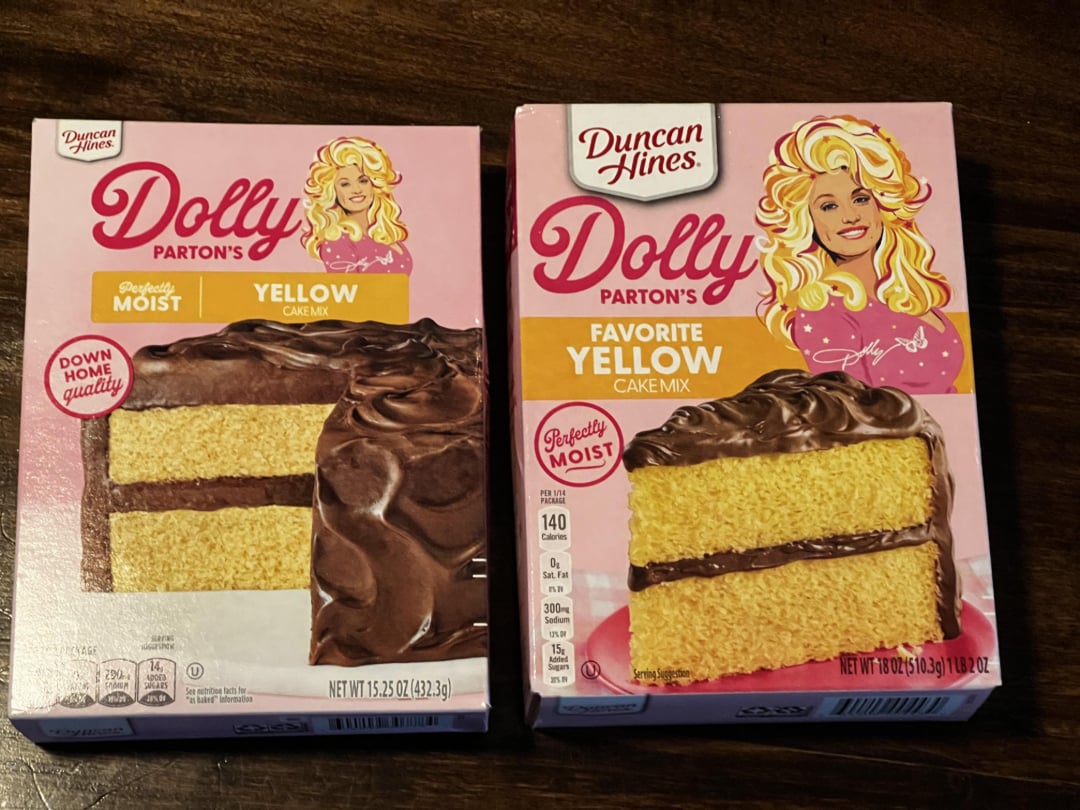 Two boxes of Duncan Hines Dolly Parton’s yellow cake mix are on a dark surface. Each box features an image of a frosted yellow layer cake and an illustrated portrait of Dolly Parton on the pink packaging.