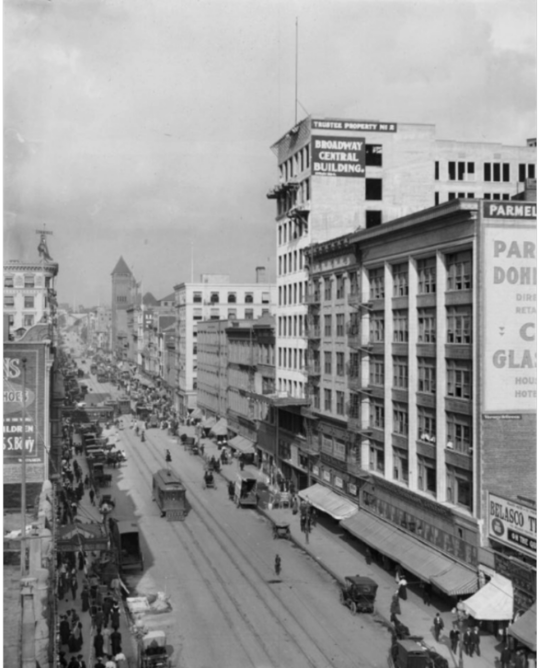 Black and white photo of a busy city street in the early 1900s, with streetcars, pedestrians, horse-drawn carriages, and multi-story buildings, including one labeled "Broadway Central Building.