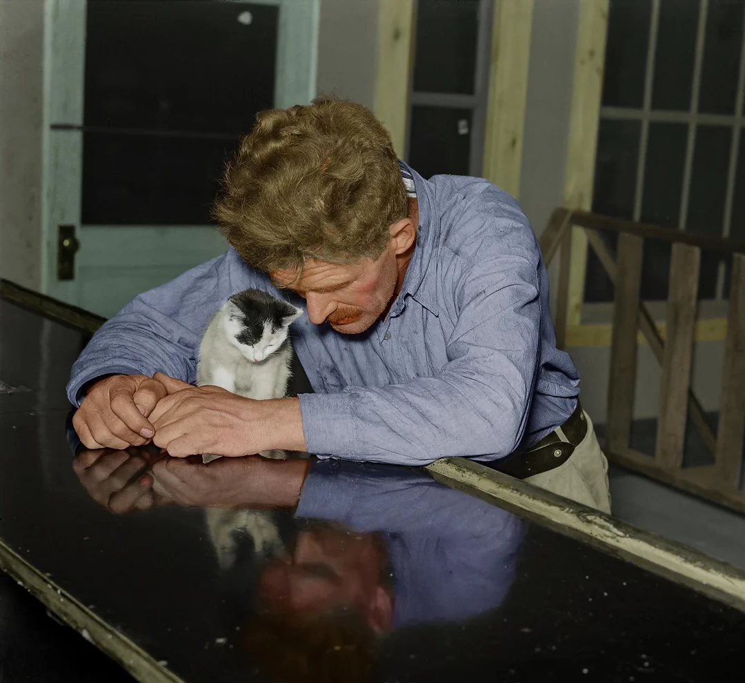 A man with wavy hair and a mustache leans on a table, bowing his head close to a small black and white cat sitting beside him, both reflected on the shiny surface of the table.