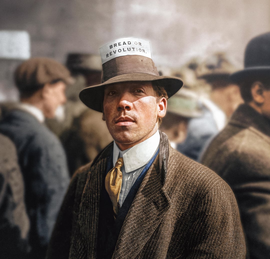 A man in early 20th-century clothing stands among a crowd, wearing a hat with a paper sign that reads "BREAD OR REVOLUTION." The scene suggests a protest or demonstration.
