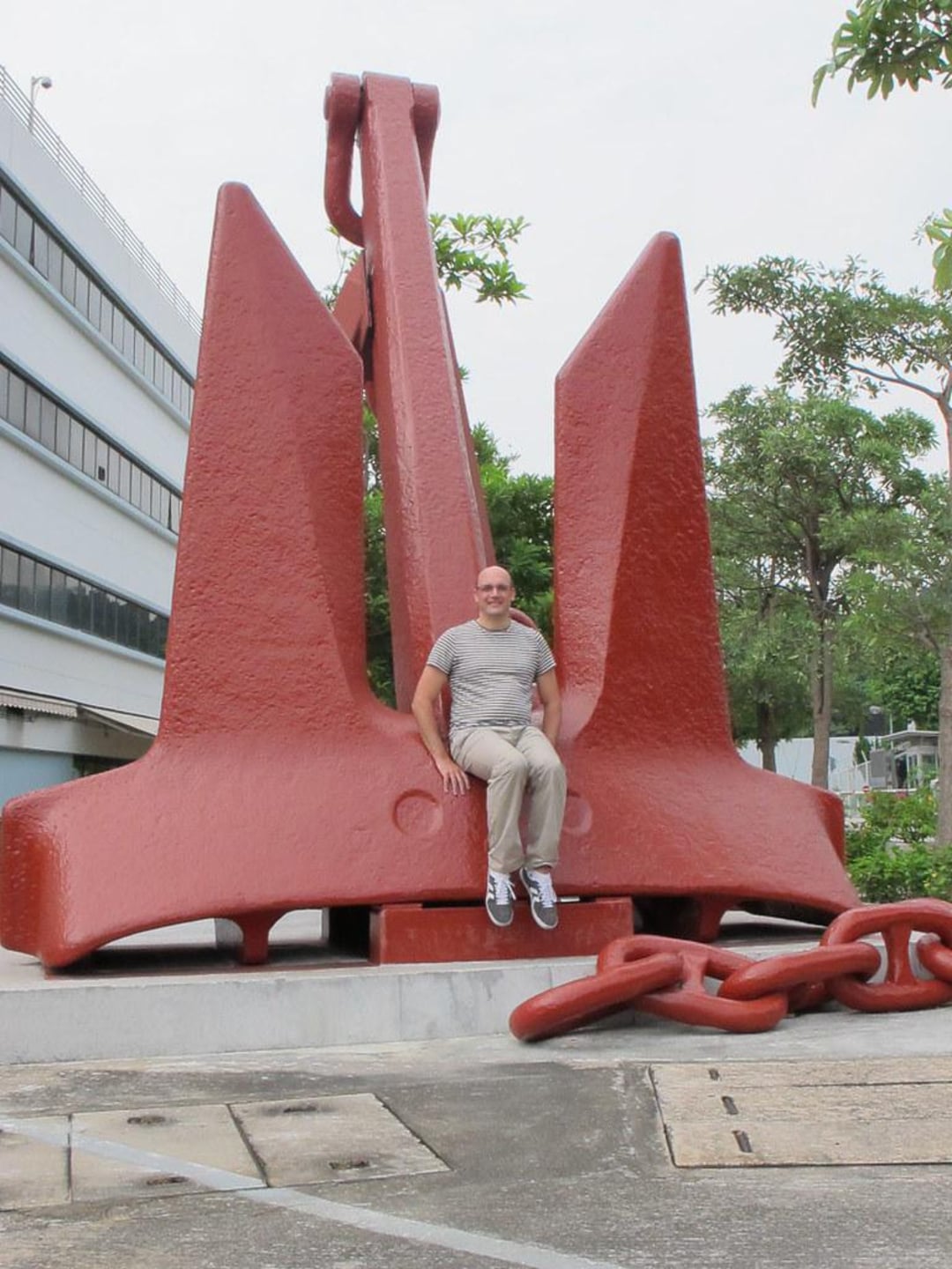 A man in casual clothing sits on the base of a massive red anchor displayed outdoors, with large chain links on the ground beside it and trees and a building in the background.