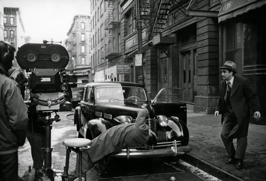 A black-and-white scene shows a film crew shooting a movie on a city street. A man in a suit stands near a vintage car while another person lies on the car’s hood. A large camera is set up to the left.