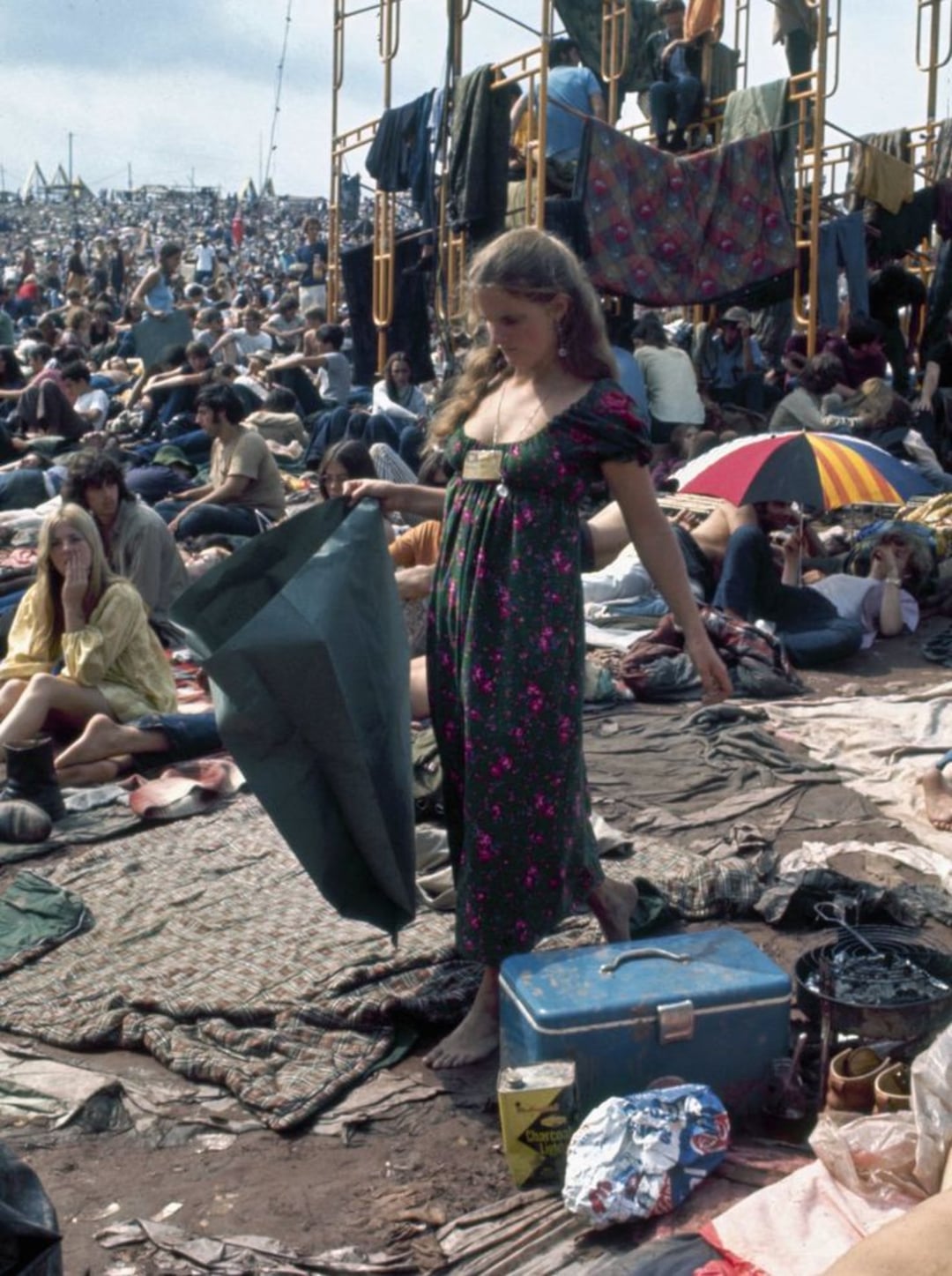 A barefoot woman in a long floral dress stands among a crowd at an outdoor festival, holding a tarp. People sit on blankets with various belongings around, and scaffolding and colorful umbrellas are visible in the background.