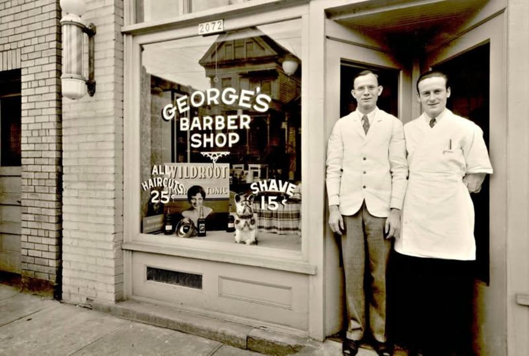 Two men in white coats stand outside "George's Barber Shop." The shop window advertises haircuts for 25¢ and shaves for 15¢. A dog sits in the window beside a photo of a man getting a haircut.