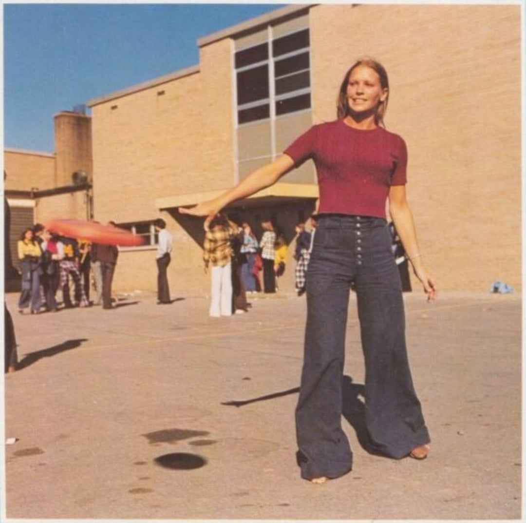 A woman wearing a red top and high-waisted bell-bottom jeans throws a frisbee outside, with a group of people and a brick building in the background.