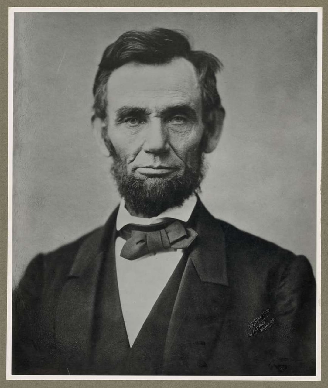 A black-and-white portrait of a man with a prominent beard, wearing a suit, bow tie, and white shirt, looking directly at the camera against a plain background.