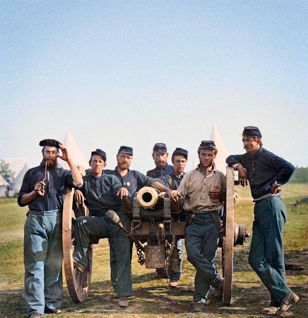 Seven Civil War soldiers in blue uniforms stand and sit around a cannon, some relaxed and some holding objects, with tents and grass in the background under a clear sky.