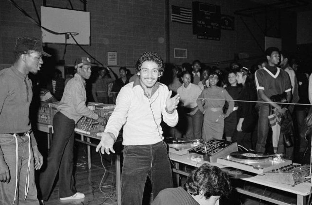 A man stands smiling and gesturing near DJ turntables at a lively indoor party, surrounded by a crowd of people enjoying the music in a gymnasium-like setting.