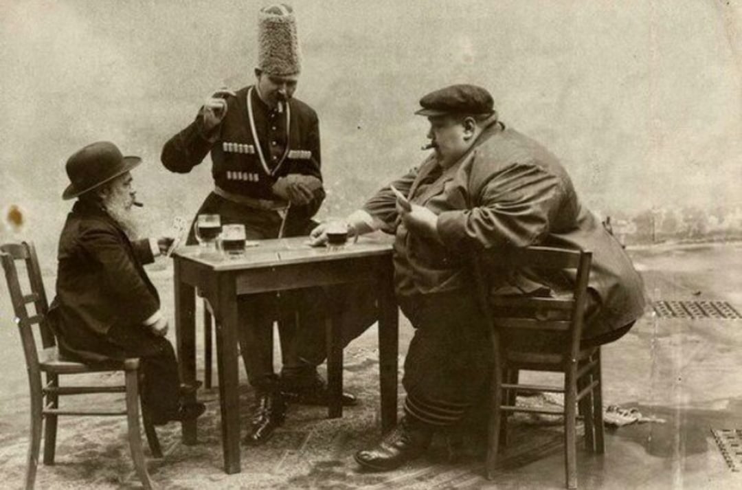 Three men sit around a small table playing backgammon; one man stands and watches, wearing traditional attire and a tall hat. The other two, differing greatly in size, are seated, with drinks and cigarettes on the table.