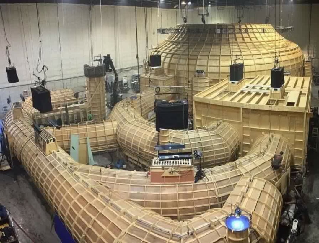 Large indoor film set with elaborate wooden structures and curved frameworks, surrounded by film equipment and lighting rigs, inside a spacious warehouse-like area. Several people are working around the set.