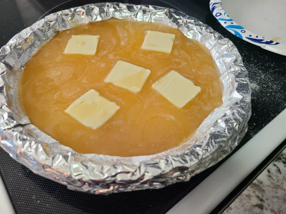 A Great Depression era pie in a foil-lined pan filled with a light brown batter, topped with five square slices of butter, sits on a stovetop next to a countertop and a paper plate.