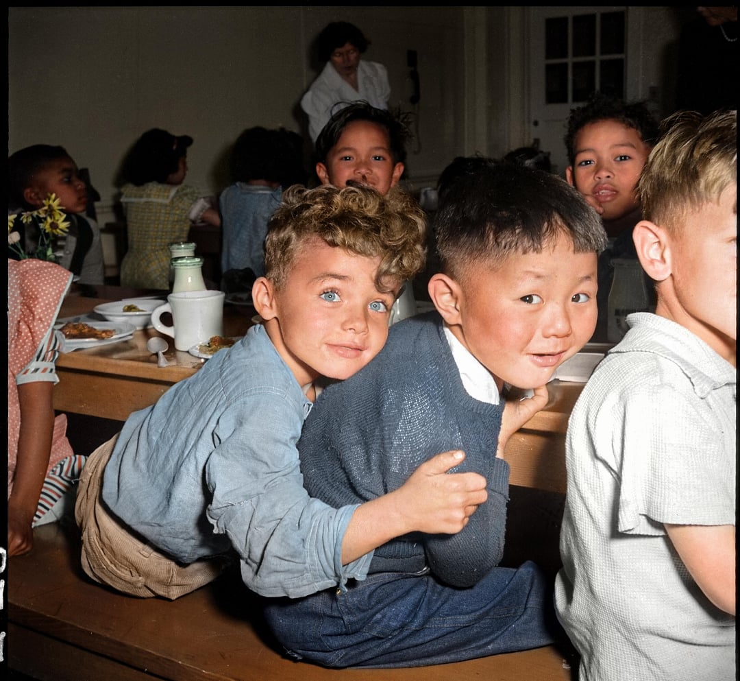 Two young boys sit closely together on a bench, smiling and looking at the camera in a busy lunchroom with other children and food trays visible in the background.
