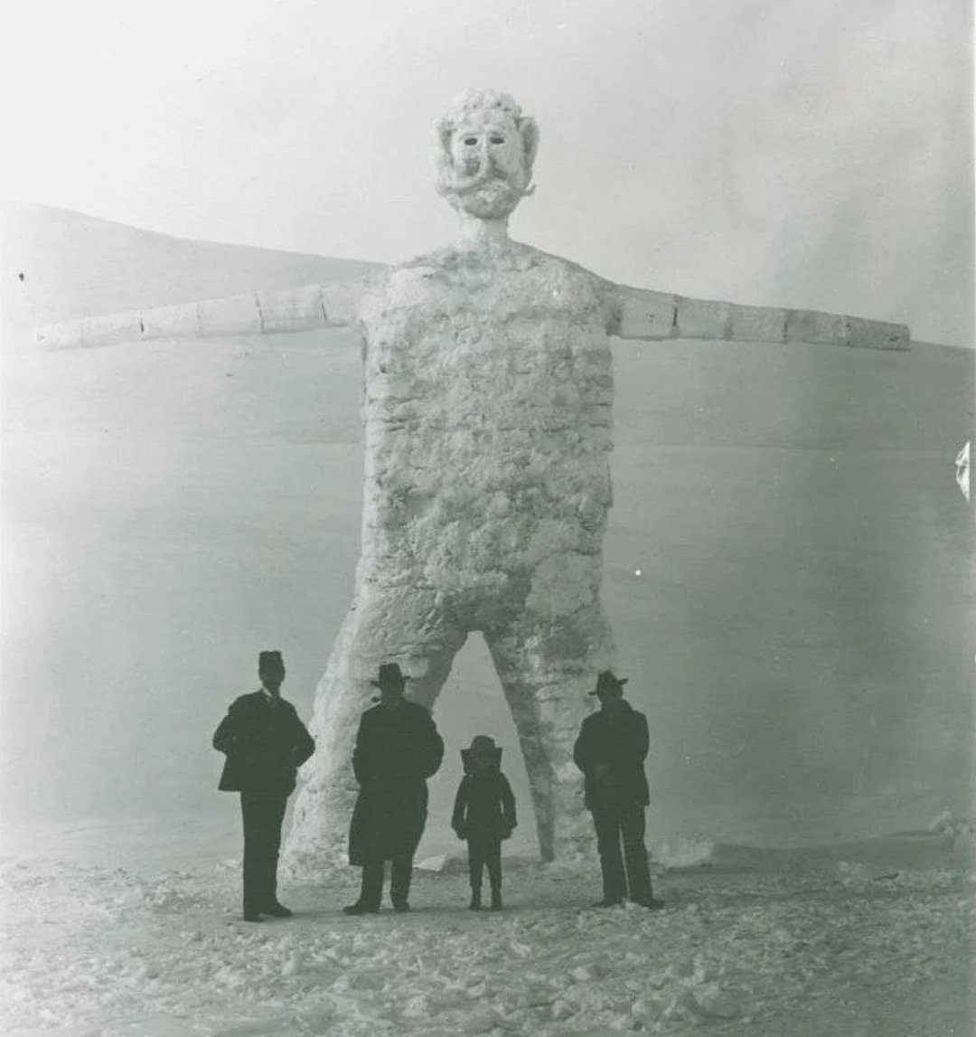 Four people stand in front of a giant snow or ice sculpture shaped like a human figure with outstretched arms, set in a snowy outdoor landscape. The scene is in black and white.