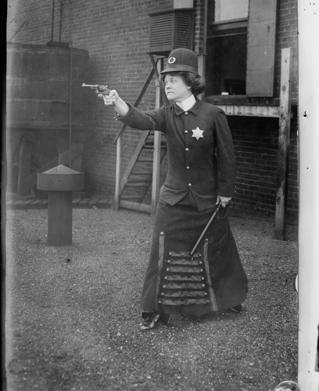 A woman in an old-fashioned police uniform and long skirt points a revolver with one hand and holds a baton in the other, standing in front of a brick building.