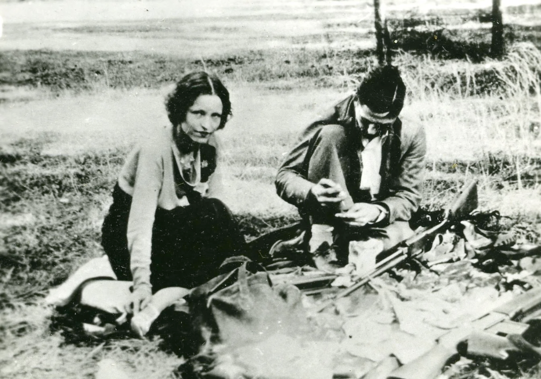 A black and white photo of a woman and a man sitting on the grass with various items laid out in front of them; the man appears to be writing or working with his hands while the woman looks toward the camera.