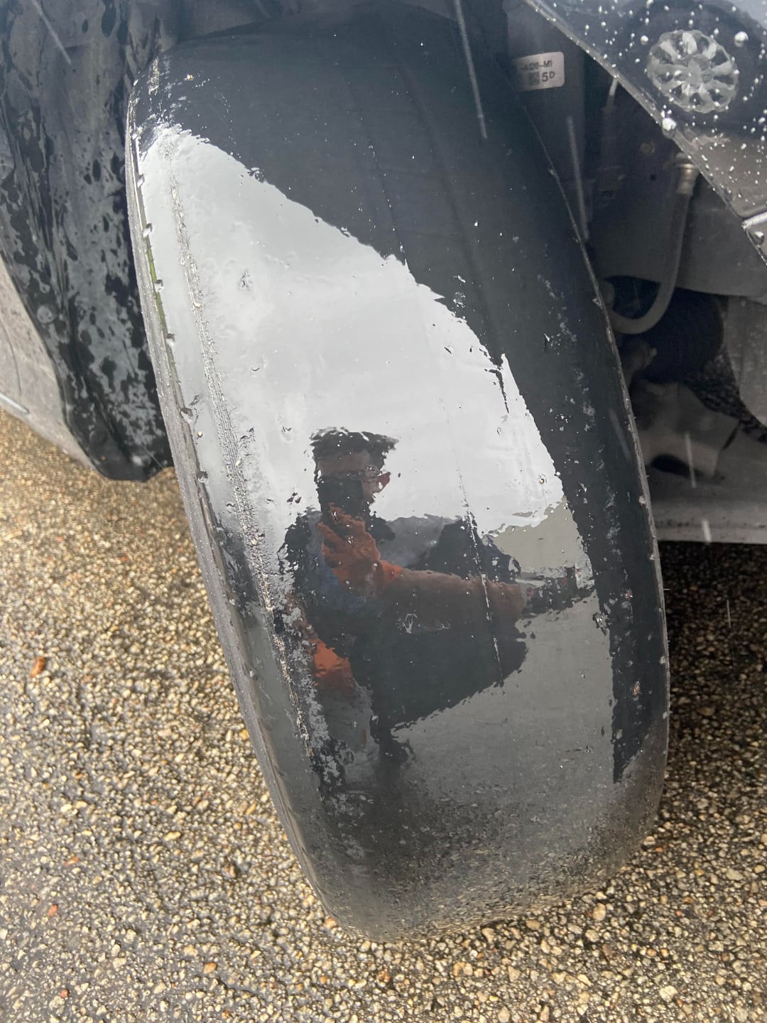A close-up of automotive fail a large, worn-out, and smooth tire with no visible tread, reflecting the image of a person, on a gravel surface.