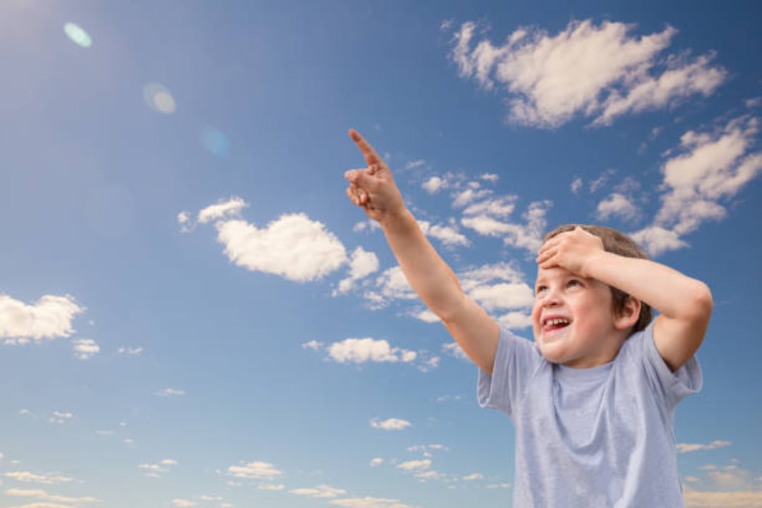 A young boy wearing a light blue shirt smiles and points excitedly at the sky with one hand, while shielding his eyes with the other, against a background of blue sky and scattered clouds.