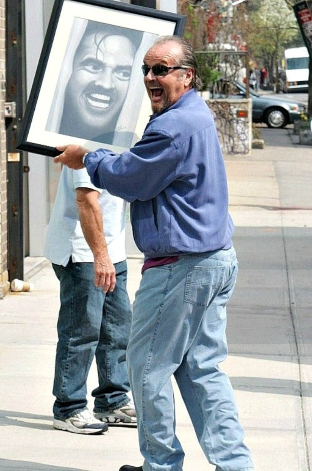 A man in sunglasses and a blue jacket happily carries a framed black-and-white portrait of a grinning face while walking on a sidewalk. Another person stands behind him, partially out of view.