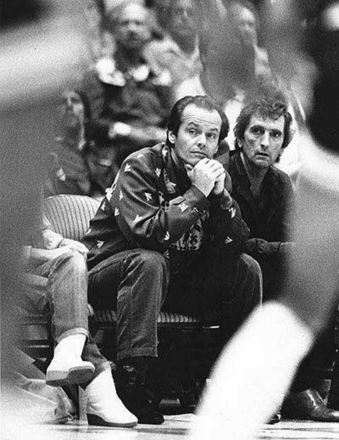 A black-and-white photo shows two men sitting courtside at a basketball game. One man leans forward with his chin resting on his hands, intently watching the action. Spectators fill the blurred background.