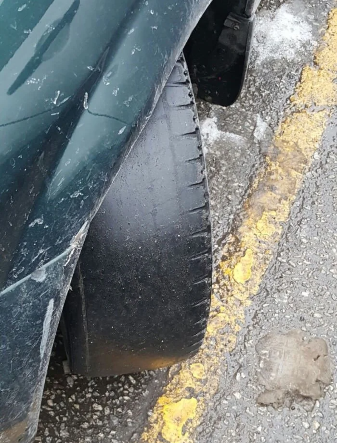 A car with a severely worn tire, showing almost no tread, parked next to a yellow line on a rough, partially snowy road.