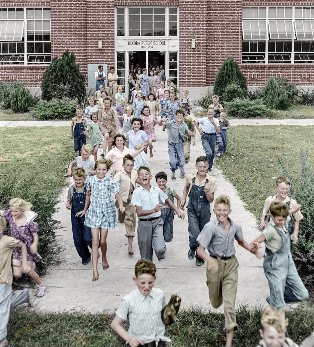 A large group of schoolchildren joyfully run out of a brick school building, smiling and laughing on a sunny day. The scene is lively, with children of various ages dressed in vintage clothing.