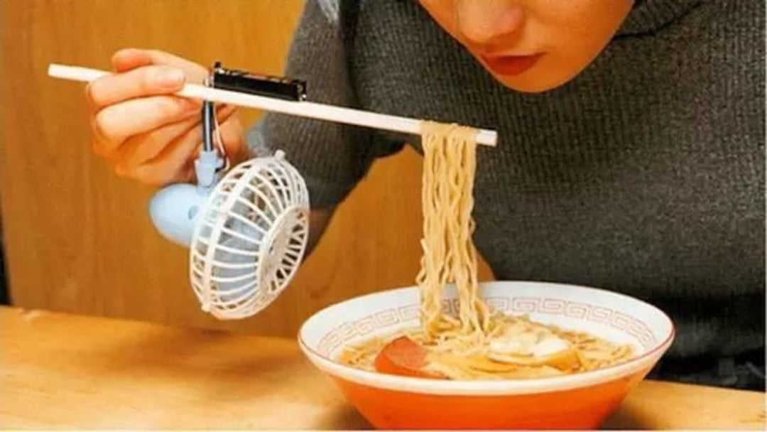 A person holds chopsticks with a small electric fan attached, cooling hot noodles above a bowl of ramen.