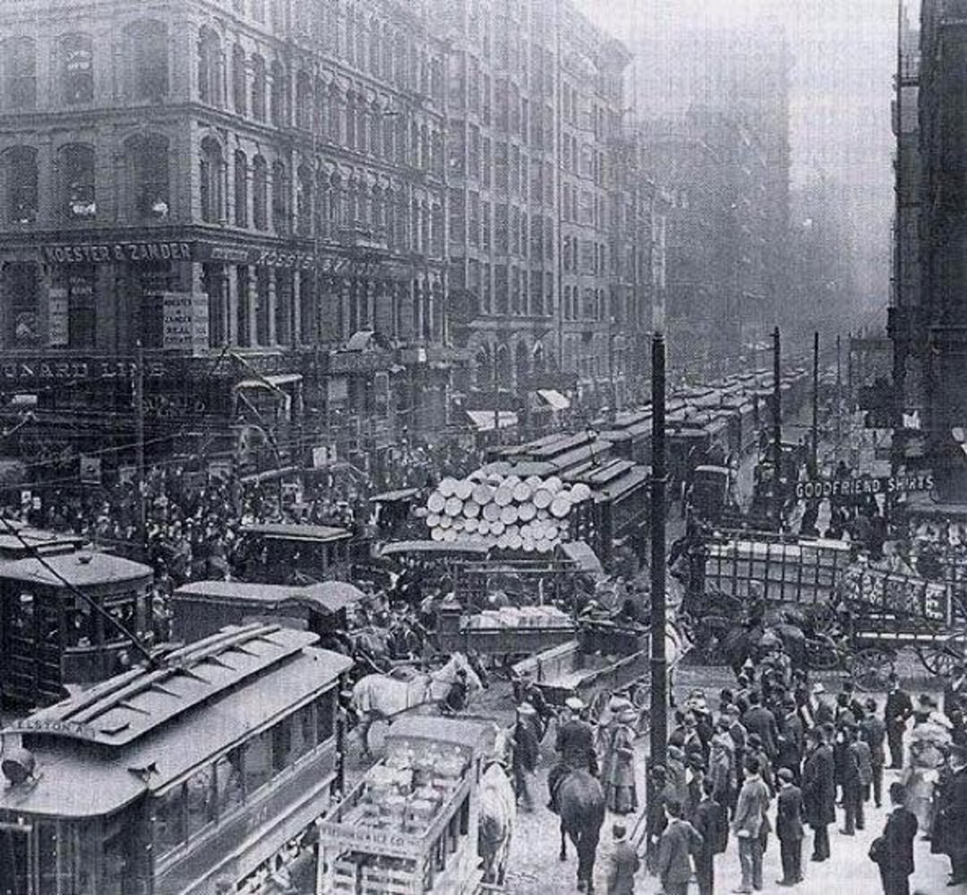 Black and white photo of a crowded city street in the early 1900s, filled with people, horse-drawn carriages, trolleys, and wagons carrying goods, surrounded by tall buildings and busy storefronts.