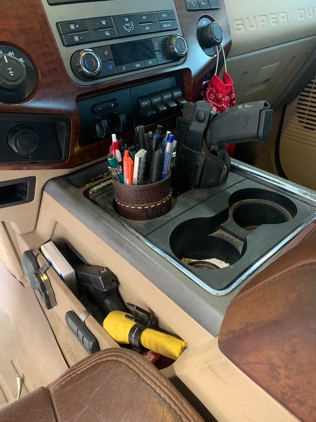 A car’s center console holding various pens in a cup, a holstered handgun, and another gun with a yellow handle tucked beside the seat. The dashboard and interior are tan and brown, with red bandanas hanging nearby.