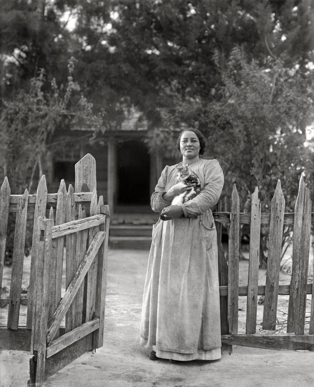 A woman stands by an open wooden gate, holding a cat in her arms. She wears a long skirt and striped jacket. Trees and a house with a porch are visible in the background.