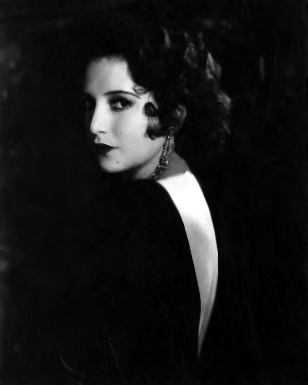 A woman with short, dark, curly hair and dramatic makeup glances over her shoulder. She wears a dark dress with a deep, open back and large, dangling earrings, set against a dark background.