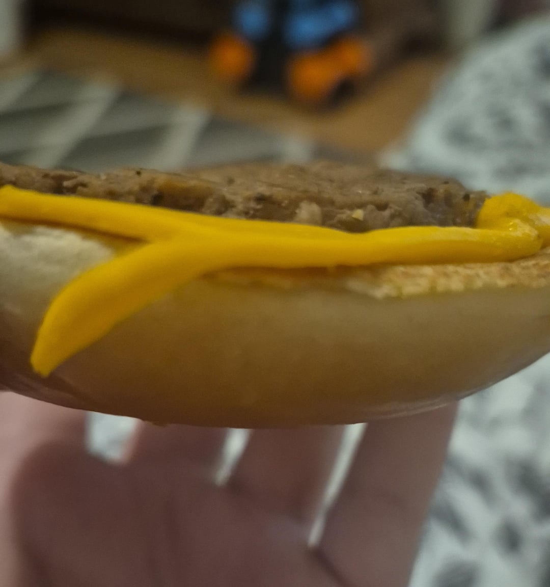 A close-up of a hand holding a hamburger with a slice of cheese unevenly placed, hanging over the edge of the bun. The background is blurred with patterned flooring and a toy visible.