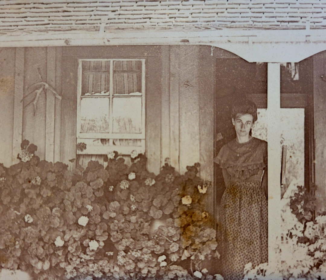 A sepia-toned vintage photo shows a woman in a patterned dress standing at the entrance of a house, surrounded by abundant flowering plants, with wooden siding and a window visible behind her.