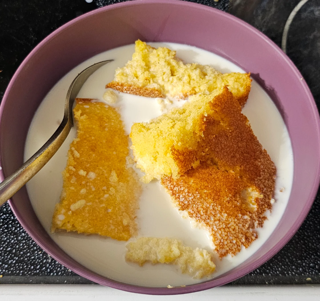 A purple bowl filled with milk and several pieces of golden-brown cornbread, with a spoon resting inside the bowl. The bowl is placed on a dark stovetop surface.