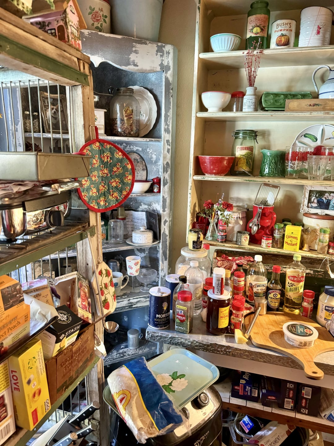 A cluttered kitchen pantry with shelves full of canned goods, jars, spices, and dishes. Countertops are crowded with condiments, bottles, and cutting boards, creating a busy, lived-in atmosphere.
