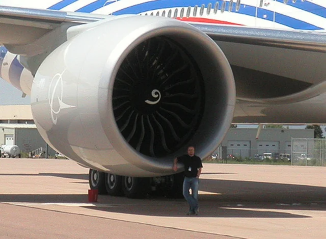 A person stands next to a very large jet engine beneath the wing of a commercial airplane, highlighting the engine's massive size compared to the individual.