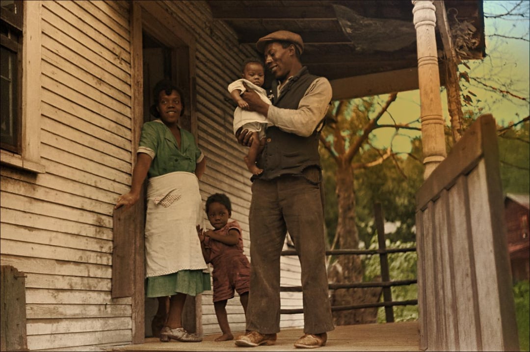 A Black family stands on the porch of a wooden house. The mother smiles by the door in a green dress and apron, the father holds a baby and wears a cap and vest, while a young child stands close to the mother’s leg.