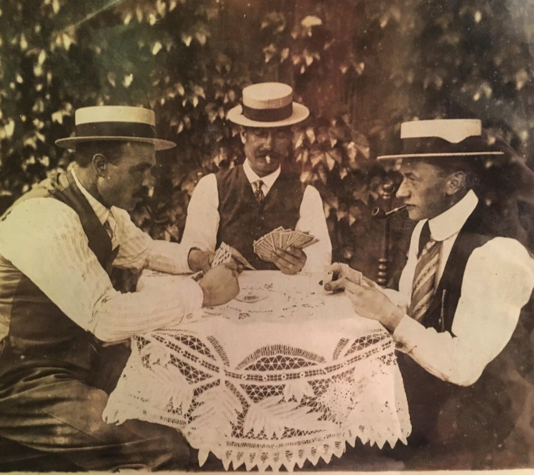 Three men in vintage clothing and straw hats sit around a lace-covered table playing cards outdoors. One holds a pipe, another deals cards, and ivy is visible in the background.