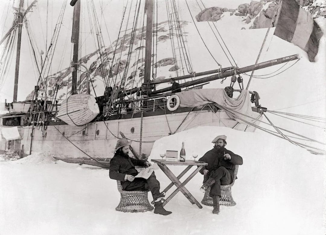 Two men sit at a small table with drinks and papers on snow, in front of a large ship trapped in ice near rocky, snow-covered terrain. The ship’s hull and ropes are visible, suggesting an Arctic or Antarctic expedition.