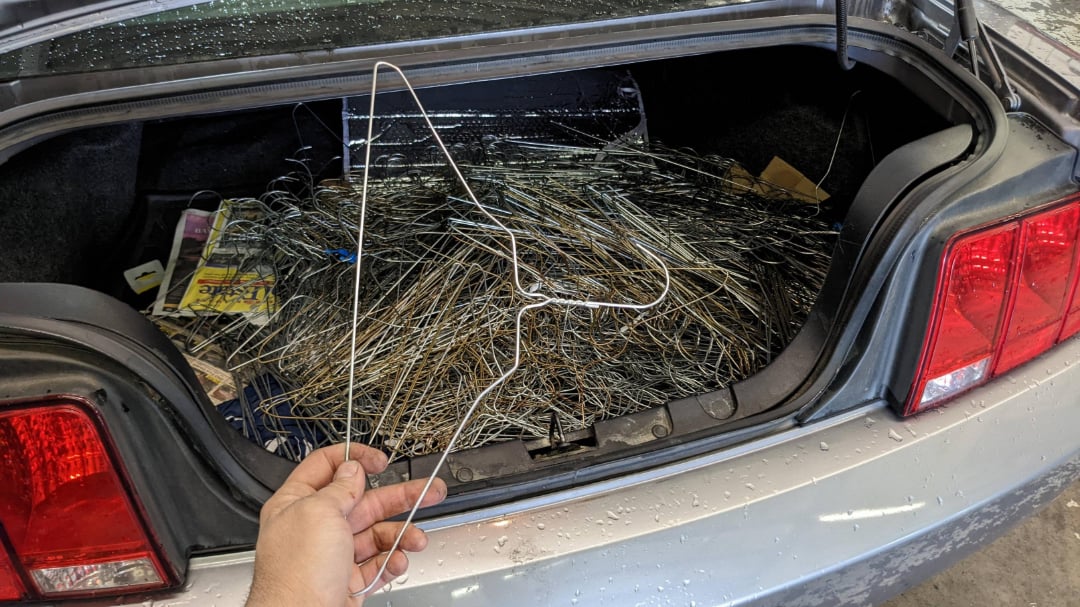 An open car trunk filled with a large pile of wire hangers. A hand is holding a single wire hanger in the foreground. Some magazines or papers are visible to the left inside the trunk.