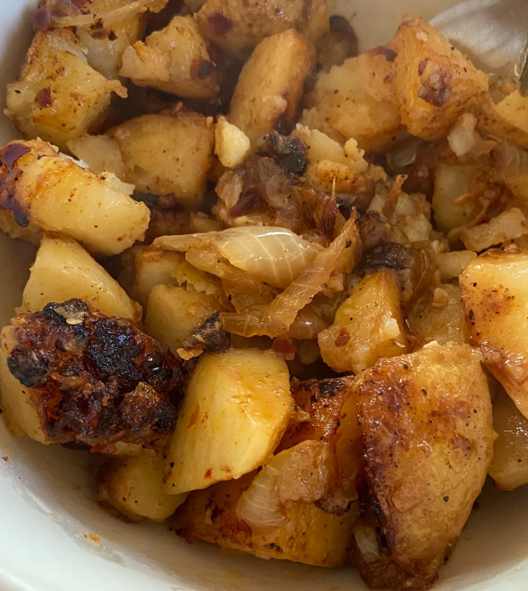 A close-up of roasted potatoes in a bowl, seasoned with spices and mixed with caramelized onions, showing crispy, browned edges and a golden-yellow color.