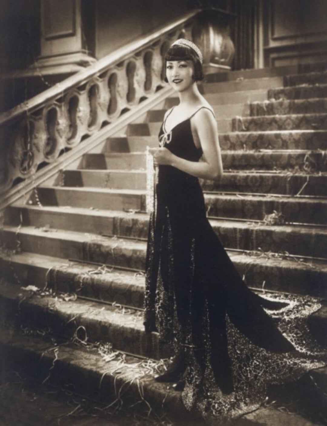 A woman in a beaded evening gown and headband stands gracefully at the base of an ornate staircase, holding a long beaded necklace. The setting and her attire evoke 1920s glamour.