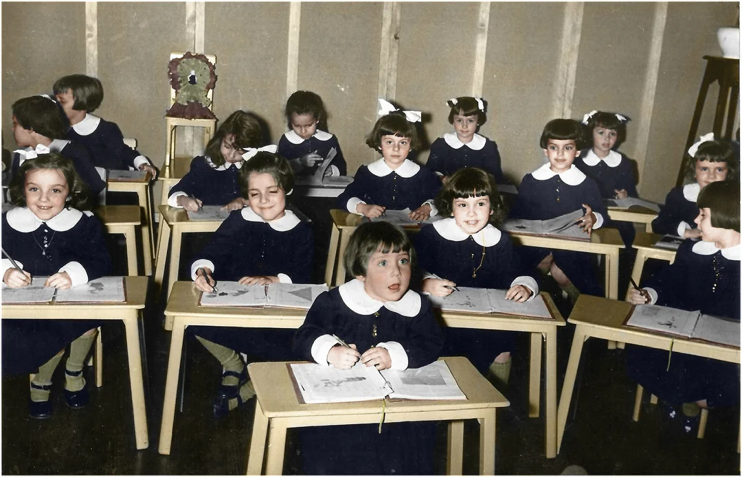 A group of young children in matching dark uniforms with white collars sit at small desks in a classroom, smiling and working in open books. The classroom has simple decor and light-colored walls.