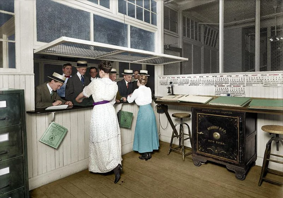 Several men in straw hats and two women in period clothing stand at a counter in an old-fashioned office with paperwork, a large safe, and filing cabinets visible in the room.