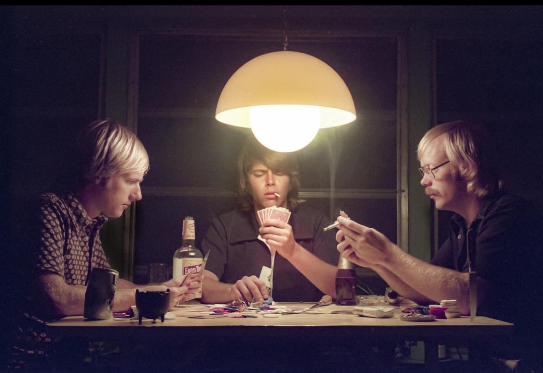 Three men sit around a table playing cards under a large hanging lamp, with poker chips, drinks, and cigarettes scattered on the table. The scene has a retro, 1970s vibe.