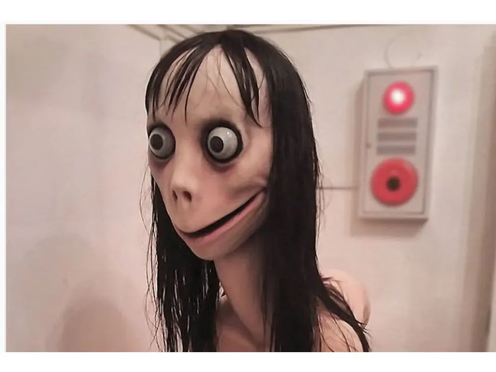 A sculpture with long black hair, bulging eyes, and a wide, unnerving smile stands in a room with white walls and a device with red lights in the background.