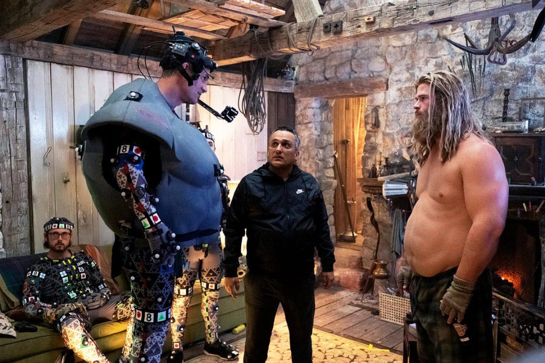 Three men stand in a rustic room. One wears a motion capture suit with a camera rig, another is in casual clothes, and the third is shirtless with long hair and gloves. Another person in a motion capture suit sits nearby.
