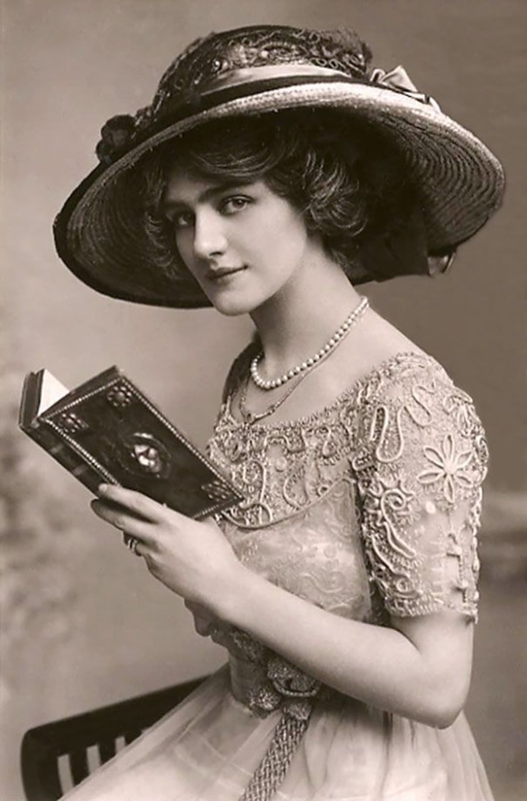 A vintage photograph of a woman in an ornate dress and wide-brimmed hat, sitting and holding an open book. She has short, wavy hair and wears a pearl necklace, looking at the camera with a slight smile.