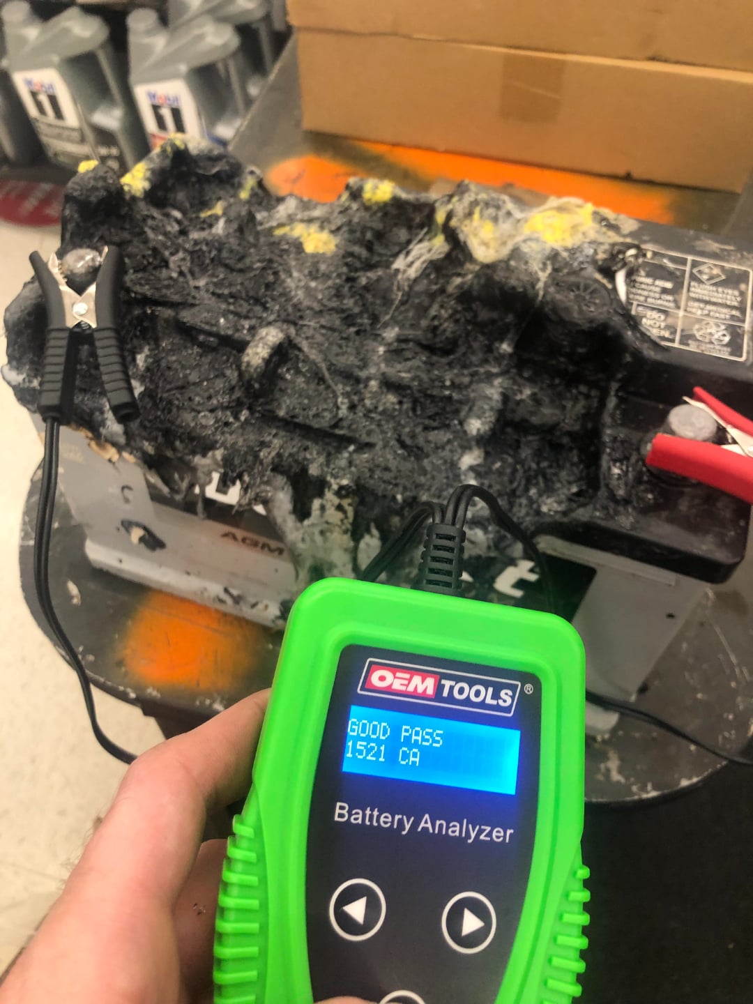 A hand holds a green battery analyzer displaying "GOOD PASS 1521 CA" near a heavily corroded and damaged car battery with attached testing clamps. Boxes and battery containers are in the background.